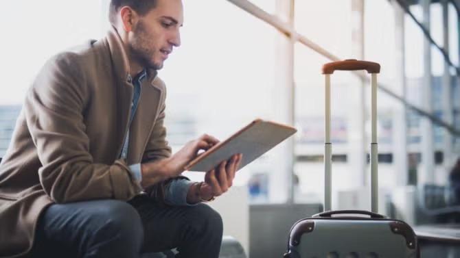 Man sitting in an airport looking at an airport with hand luggage demonstrating the benefits of packing light for business trips. 